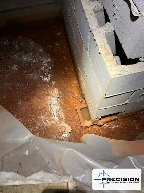 Real North Georgia crawlspace with wet exposed red clay, water pooled at the base of a concrete block foundation, and a pulled-back plastic vapor barrier
